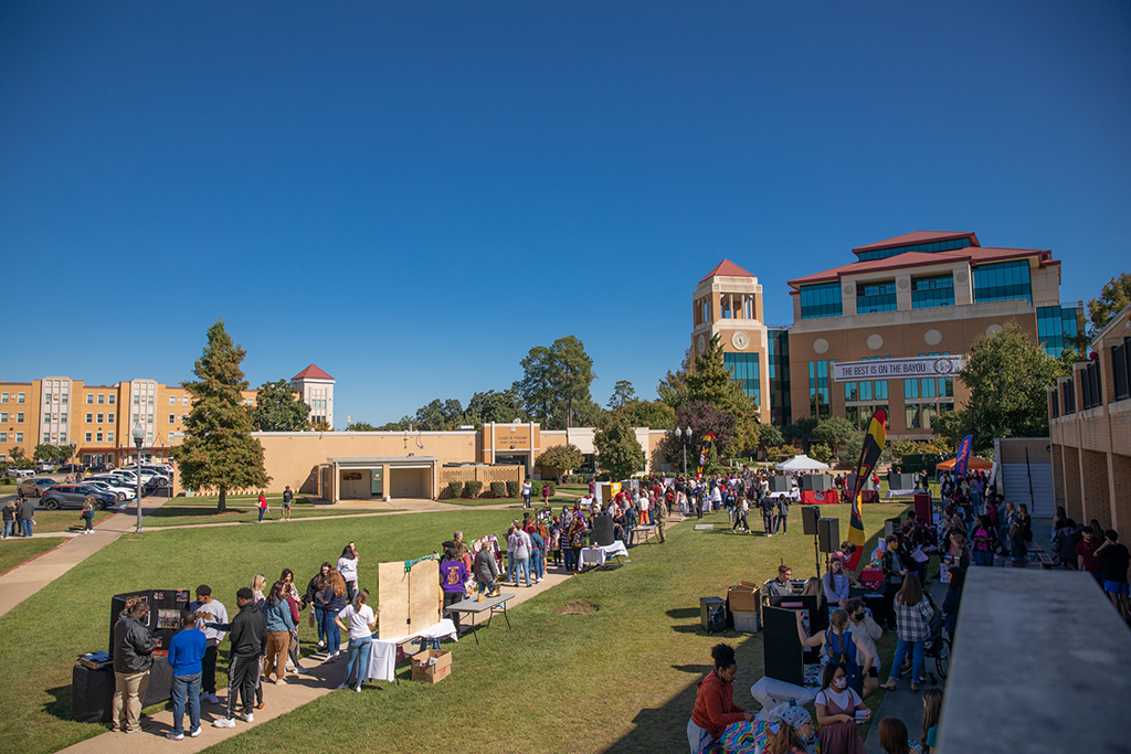 People mingle at booths and tables in the quad on 's campus. A banner hanging from the library in the background reads "The Best is on the Bayou."